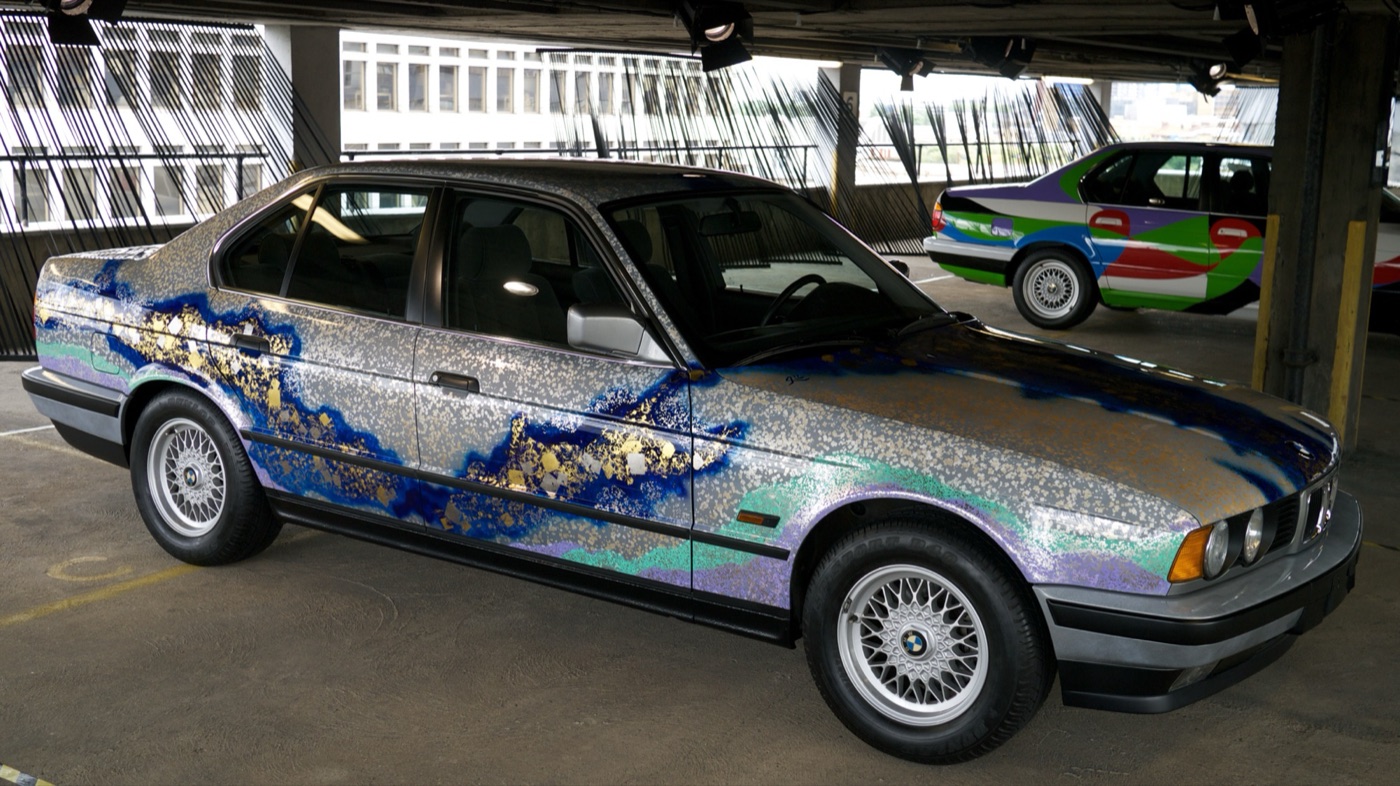 Matazo Kayama’s silver BMW 535i Art Car with blue and gold foil patterns along its side, parked in a multi-storey car park, with César Manrique’s 730i behind it