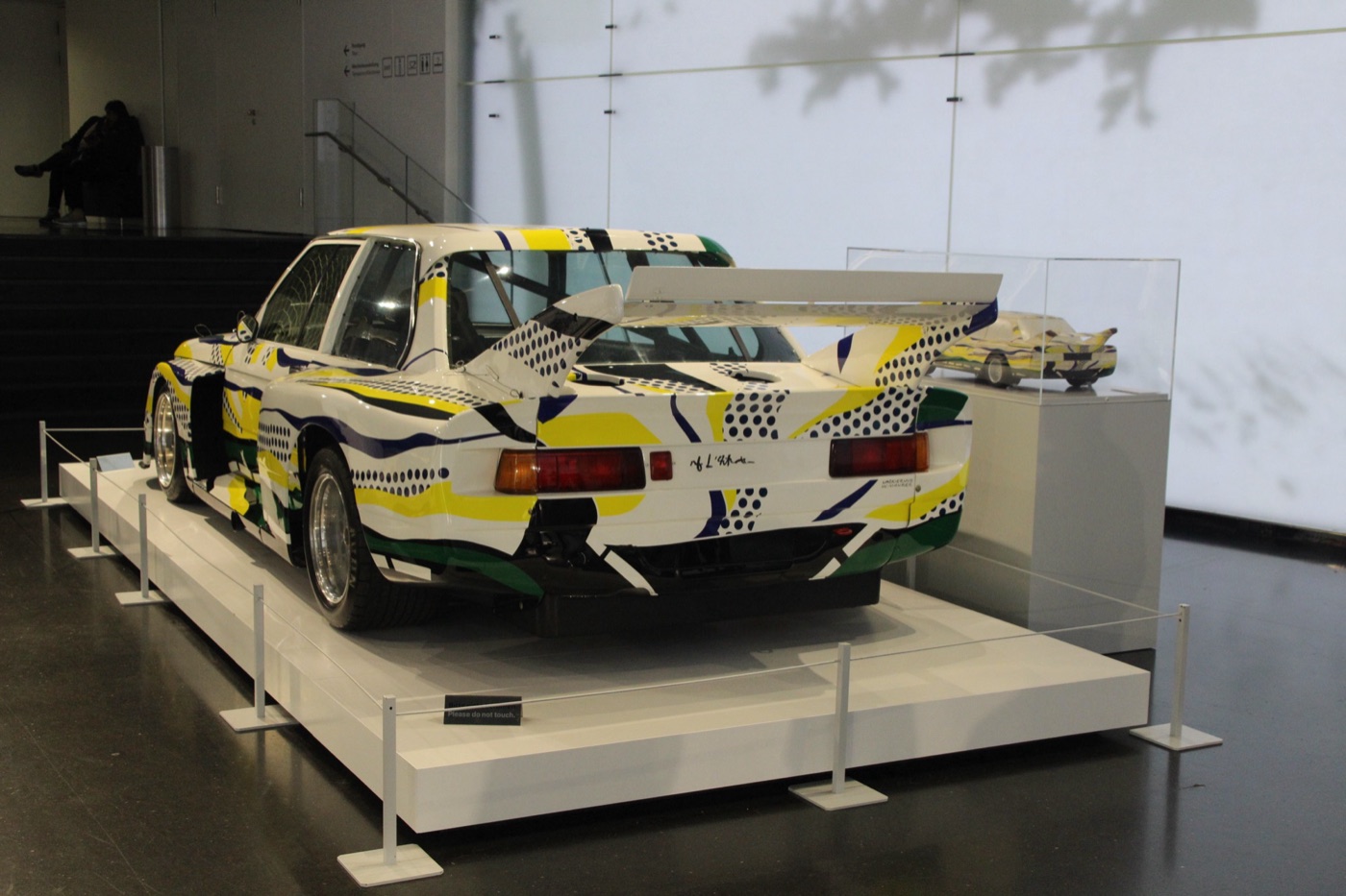 Rear three-quarter view of Roy Lichtenstein’s BMW 320 Art Car with yellow, green and blue stripes and black dots, a large rear wing and Lichtenstein’s signature above the rear lights