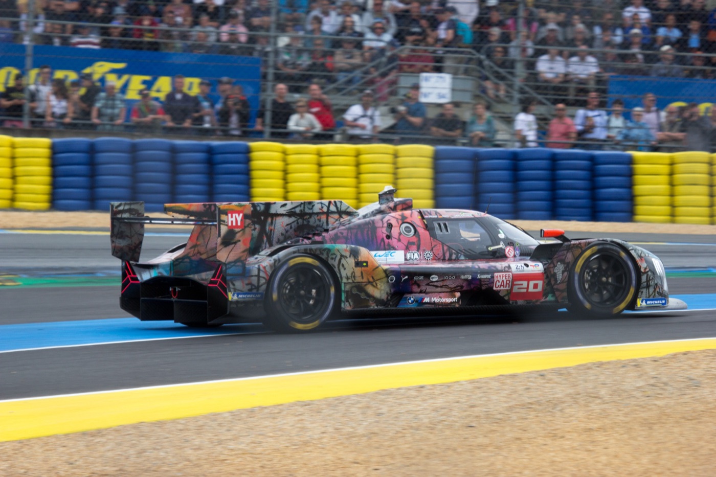 Julie Mehretu’s BMW M Hybrid V8 Art Car, race number 20, in layered pink, black and grey markings, at speed past a tyre wall and a grandstand at Le Mans
