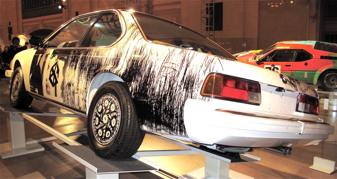 Robert Rauschenberg’s white BMW 635 CSi Art Car from the rear three-quarter, its side and boot covered in black-and-white photographs, on a display stand with Calder’s 3.0 CSL behind