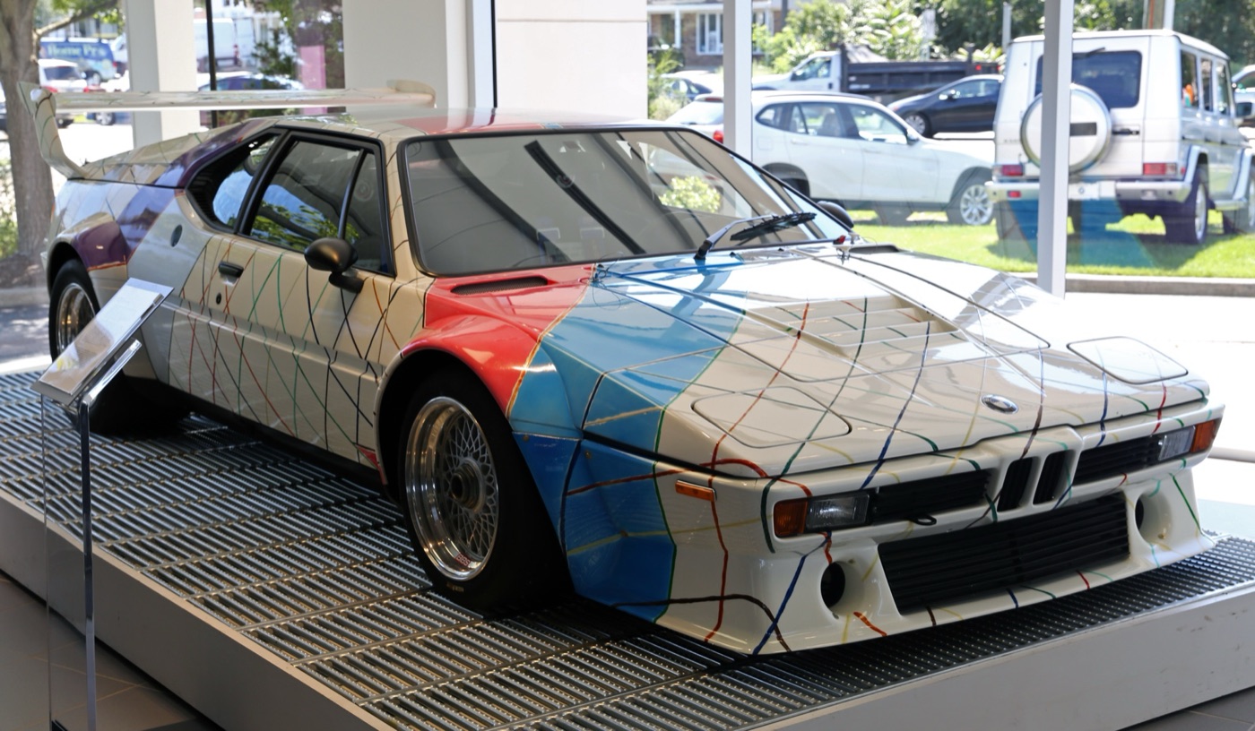 White BMW M1 Procar painted by Frank Stella with a grid of coloured lines and blocks of red and blue, on a display platform in a showroom