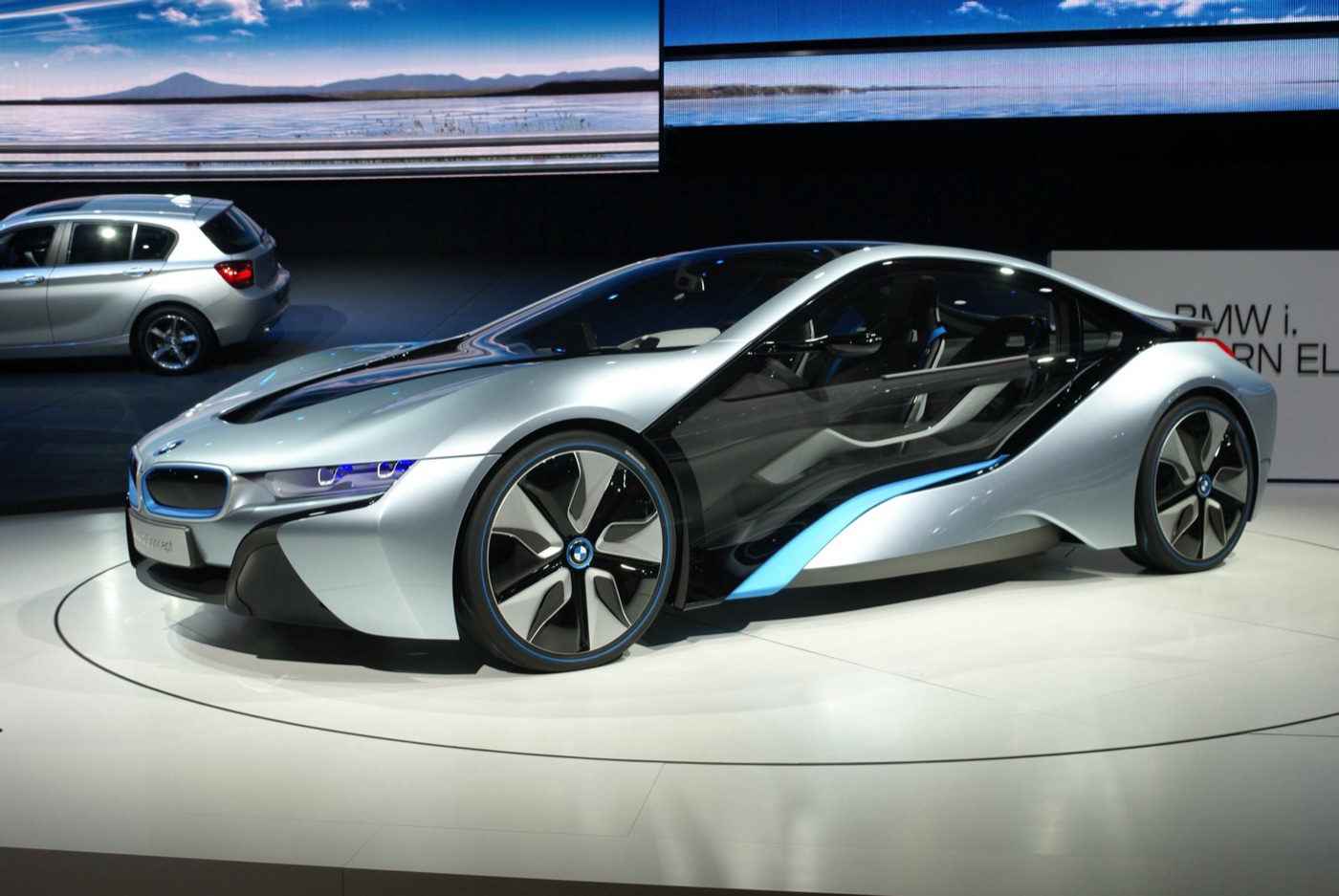 Silver BMW i8 Concept with transparent doors and turbine-style wheels on a white turntable, a BMW 1 Series behind
