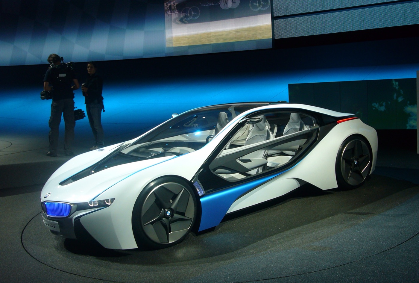 White and blue BMW Vision EfficientDynamics concept with glass doors and blue-lit kidney grille on a turntable, a cameraman behind it