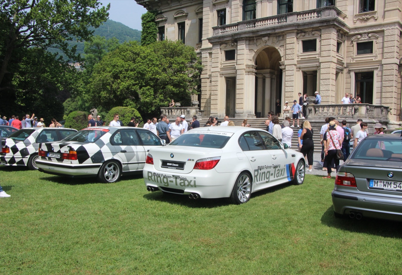 White E60 BMW M5 in BMW Driver Training Ring-Taxi livery with M stripes, parked on a lawn beside two older M5s in black and white chequered BMW M livery