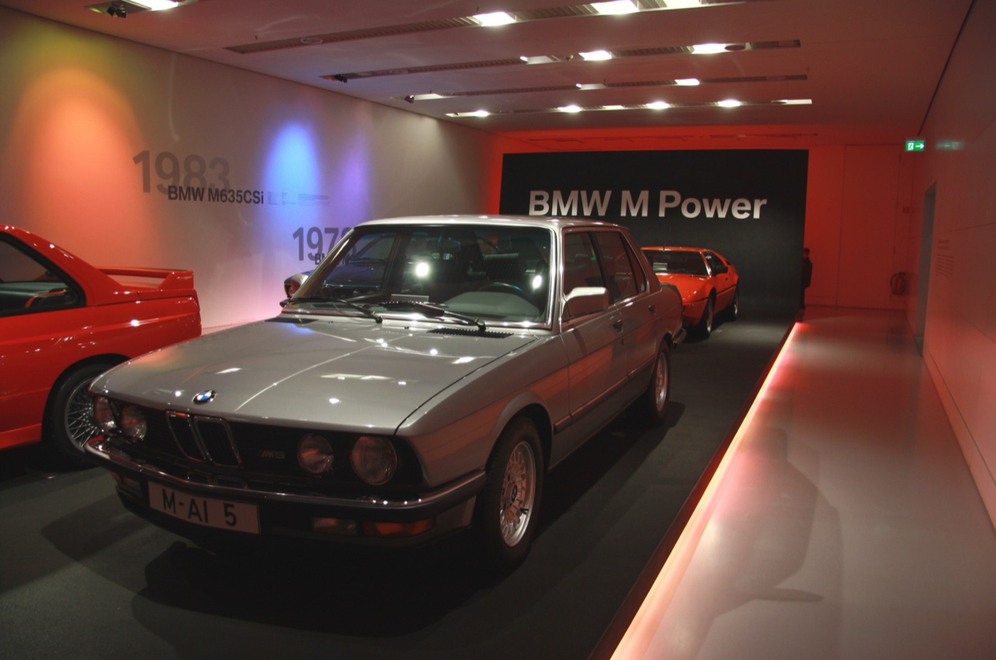 Silver first-generation BMW M5 saloon on display in front of a BMW M Power sign, with a red E30 M3 to the left and an orange M1 behind