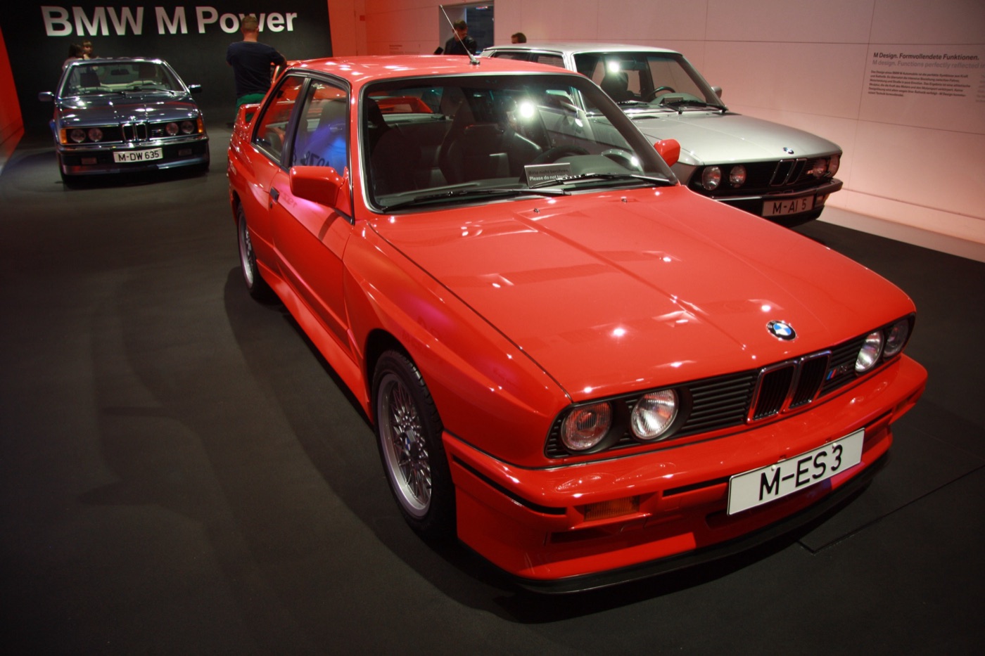 Red E30 BMW M3 Sport Evolution on display under a BMW M Power sign, with a dark blue M635CSi and a silver E28 M5 behind it