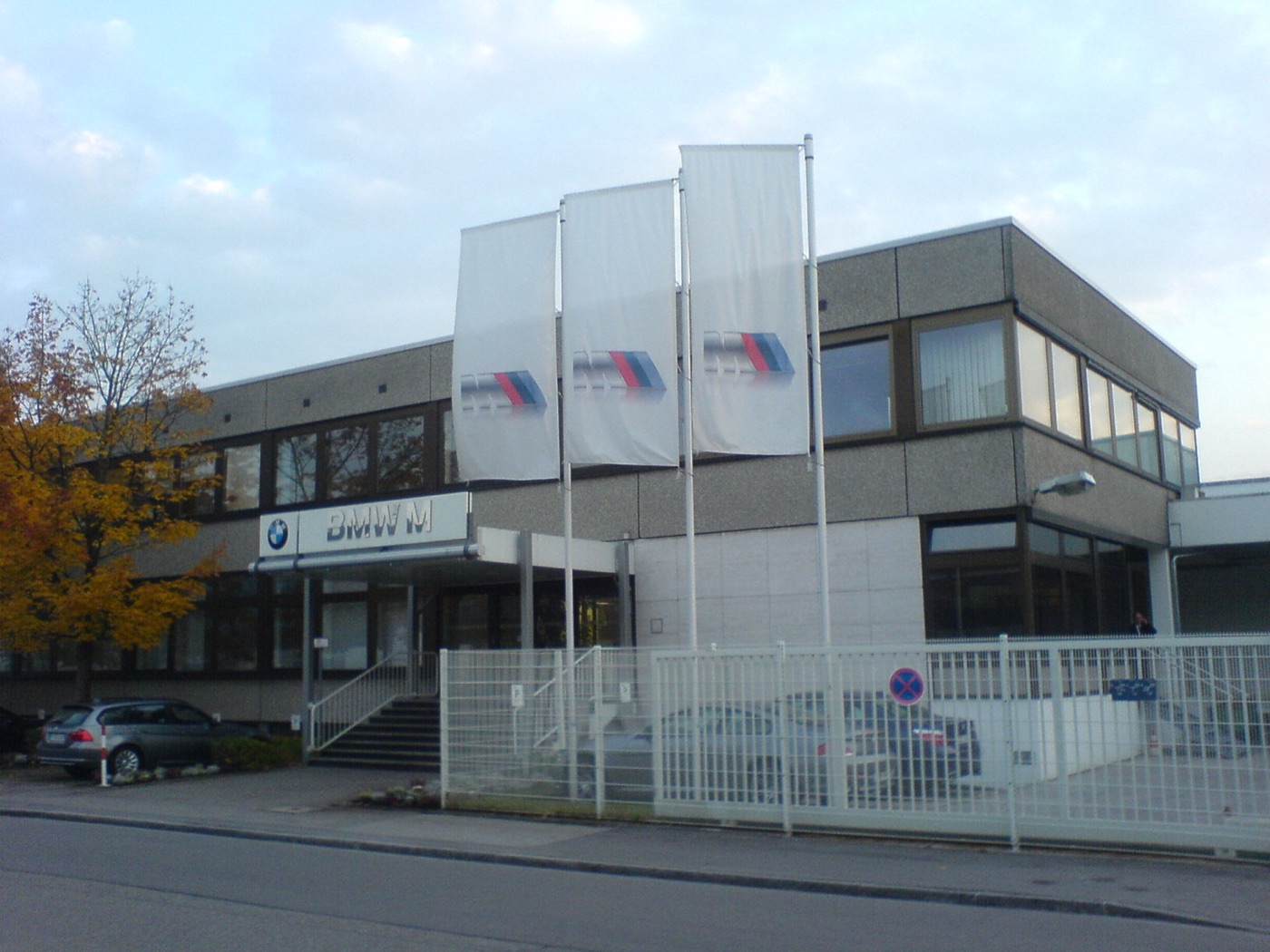 Two-storey grey office building with a BMW M sign over the entrance and three white flags carrying the M logo, behind a white steel fence