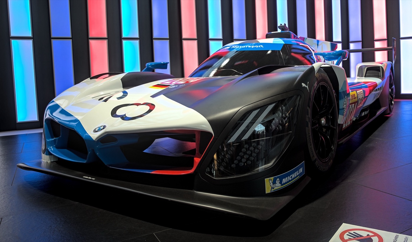 BMW M Hybrid V8 endurance prototype in white, blue and black with M Motorsport on the windscreen strip, displayed in front of light panels in the blue, dark blue and red M colours