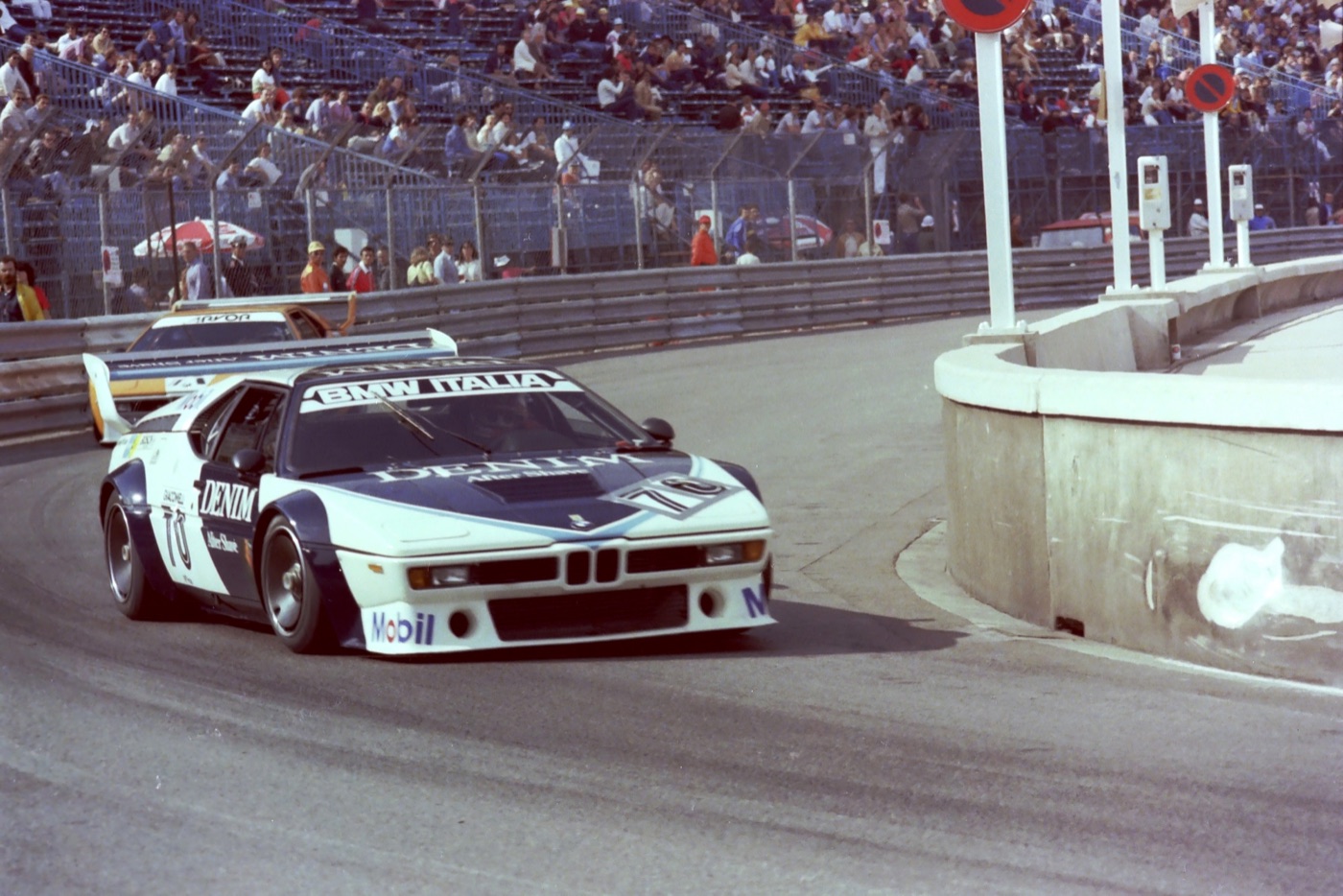 White BMW M1 Procar racing car with dark blue Denim sponsorship and BMW Italia on the windscreen, number 70, cornering at Monaco in front of a full grandstand