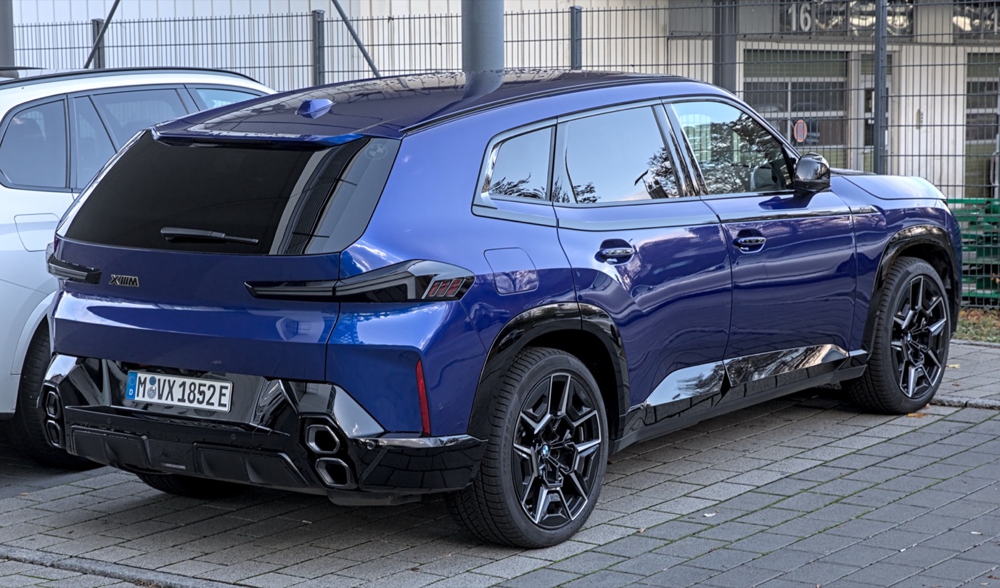 Blue BMW XM plug-in hybrid SUV from the rear three-quarter, with XM badge, black lower bodywork and stacked hexagonal exhaust tips, parked on paving