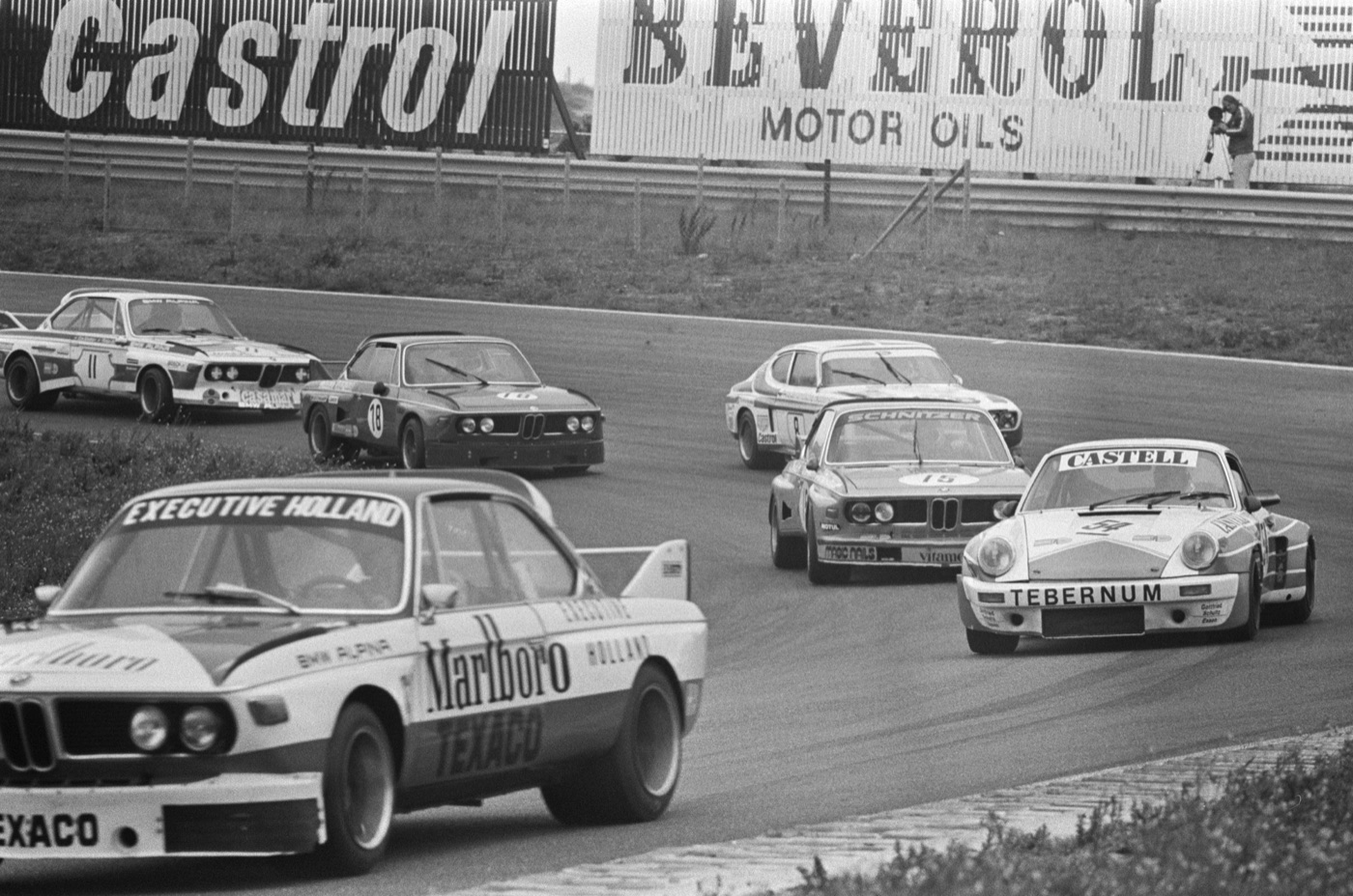 Black and white photograph of several BMW 3.0 CSL coupes, one with a large rear wing and Texaco lettering, racing with a Porsche 911 and a Ford Capri round a bend at Zandvoort