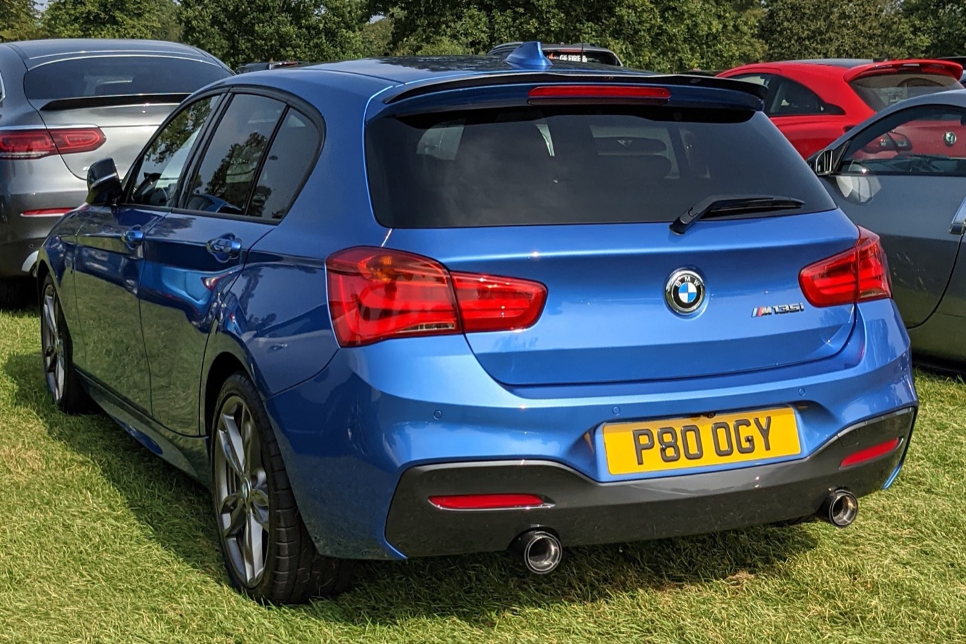 Rear of a blue facelifted BMW M135i five-door with L-shaped tail lights, M135i badge and twin exhaust pipes, parked on grass