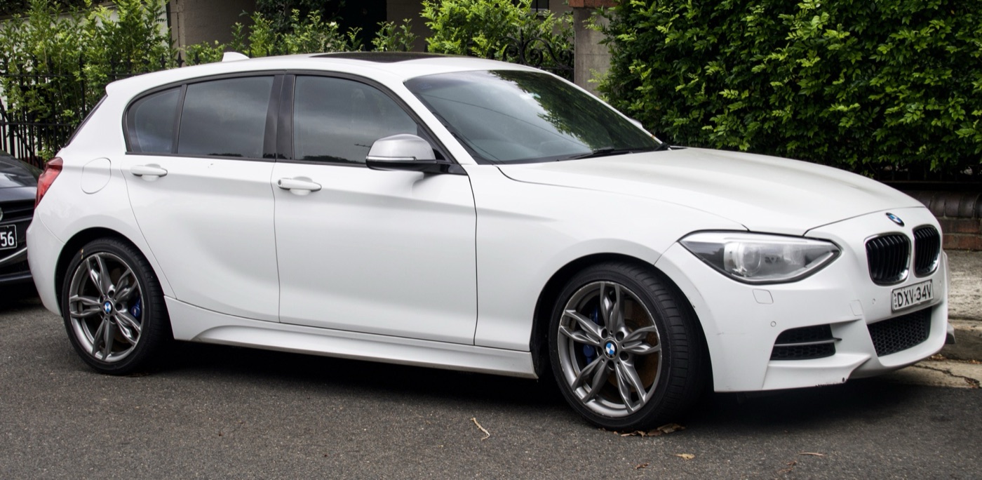 White BMW M135i five-door with grey M double-spoke alloy wheels and blue brake calipers parked at a kerb in front of a hedge