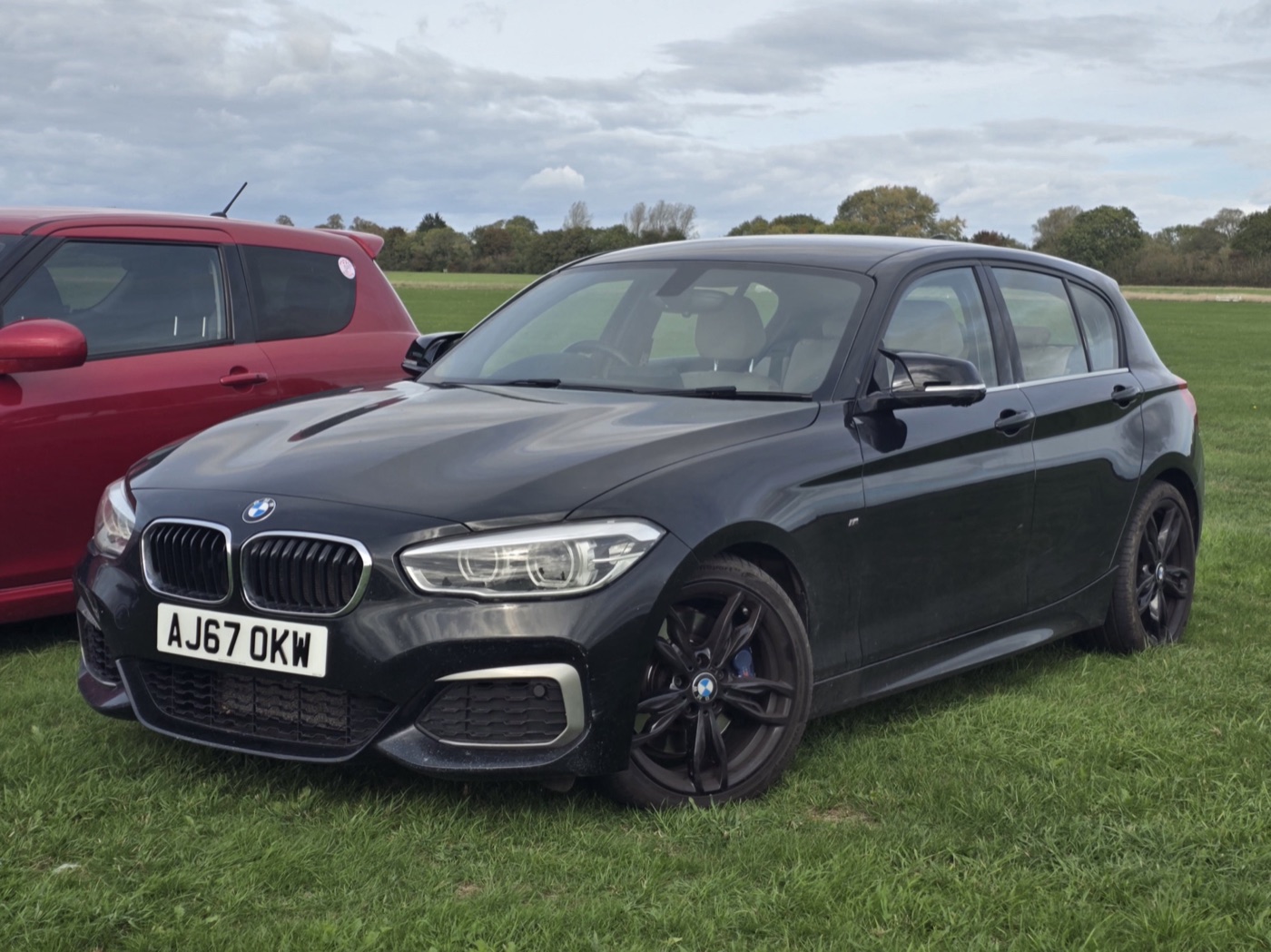 Black BMW M140i five-door with dark alloy wheels and LED headlights, parked on grass beside a red hatchback