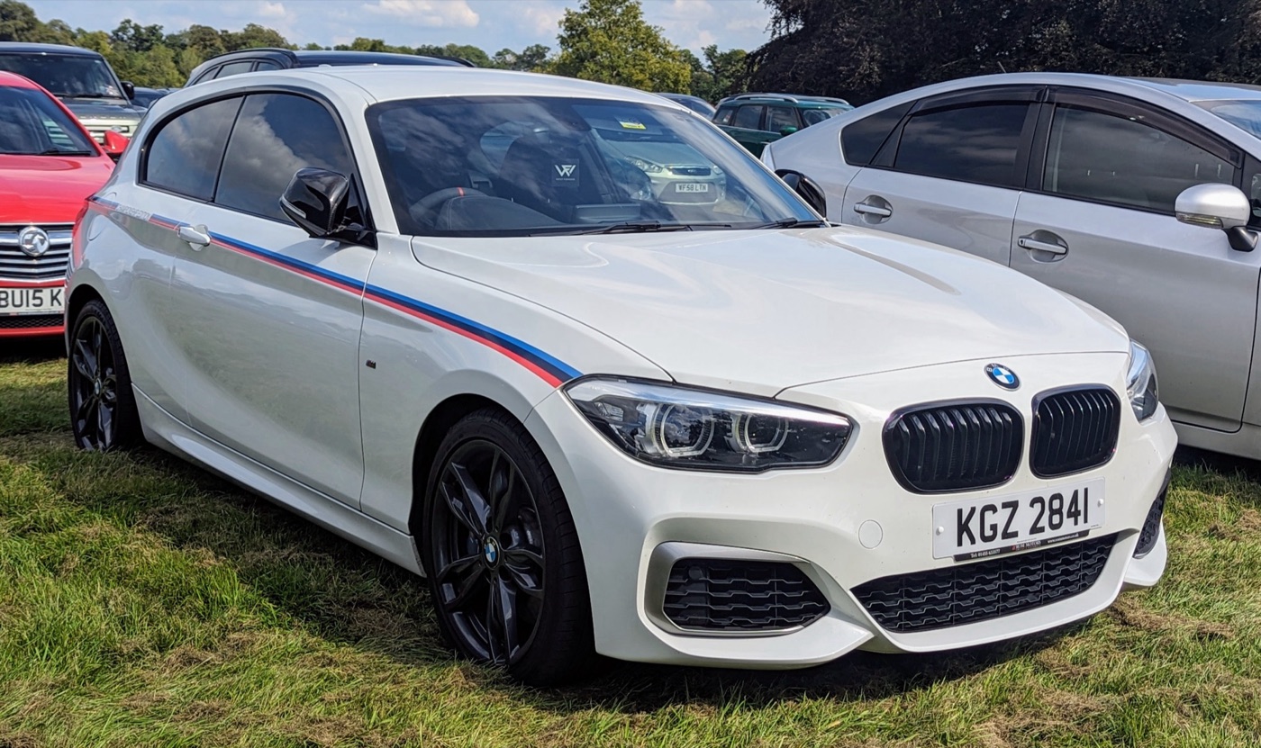 White BMW M140i Championship Edition three-door with blue, violet and red stripes along its side and black alloy wheels, parked on grass