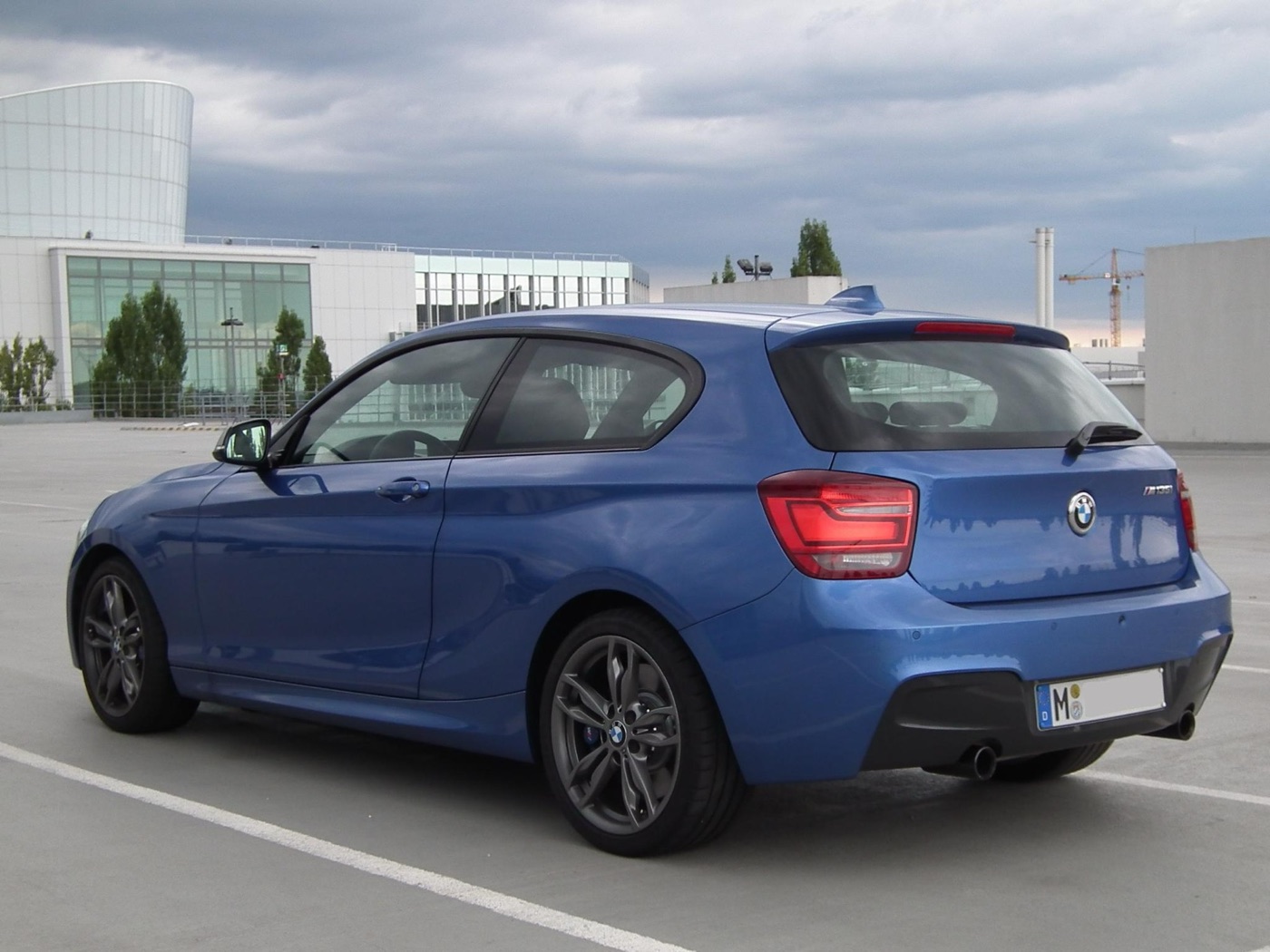 Blue BMW M135i three-door seen from the rear three-quarter on a rooftop car park, with twin exhaust pipes and grey alloy wheels