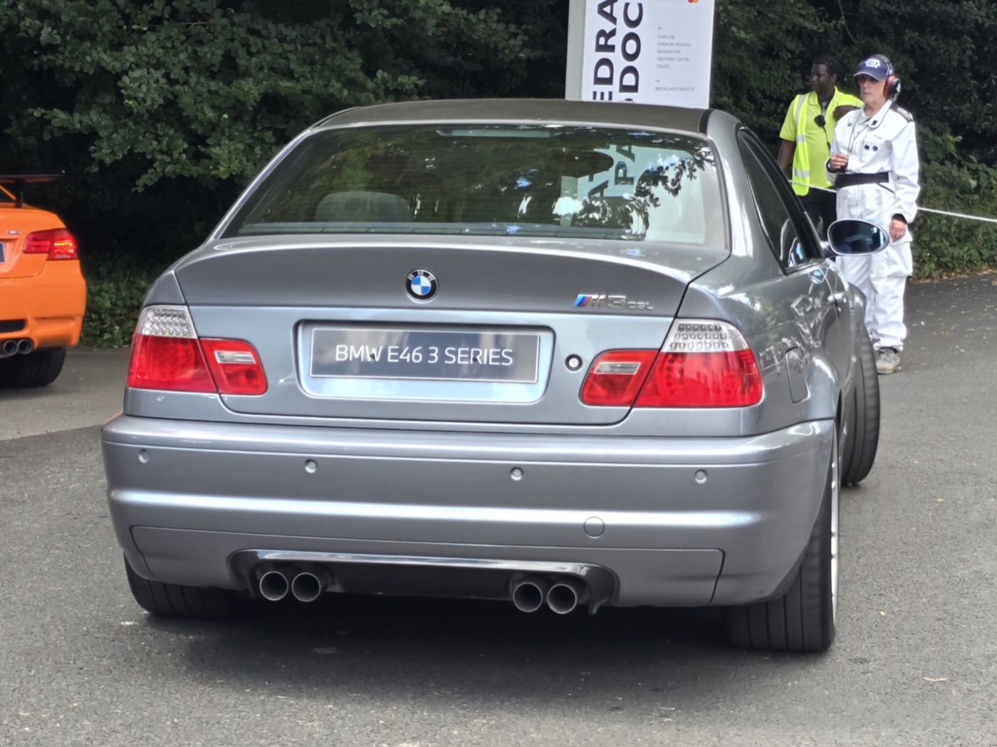 Grey E46 BMW M3 CSL from the rear, showing the M3 CSL badge and four exhaust tips, with an orange M3 GTS at the left edge