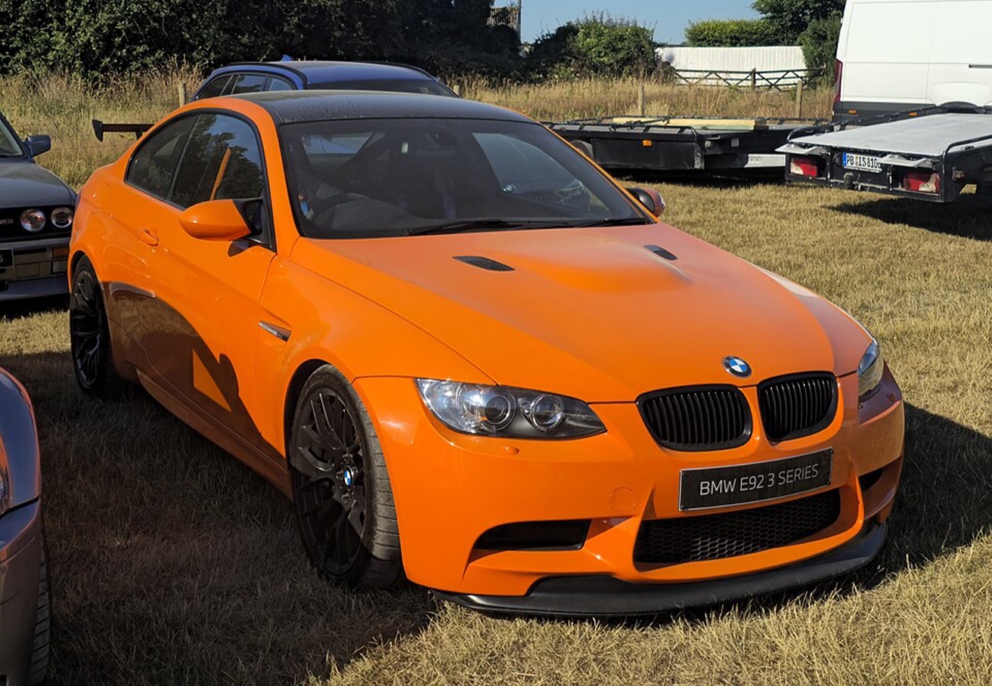 Fire Orange BMW M3 GTS from the front three-quarter, with black wheels, carbon roof and front splitter, parked on grass