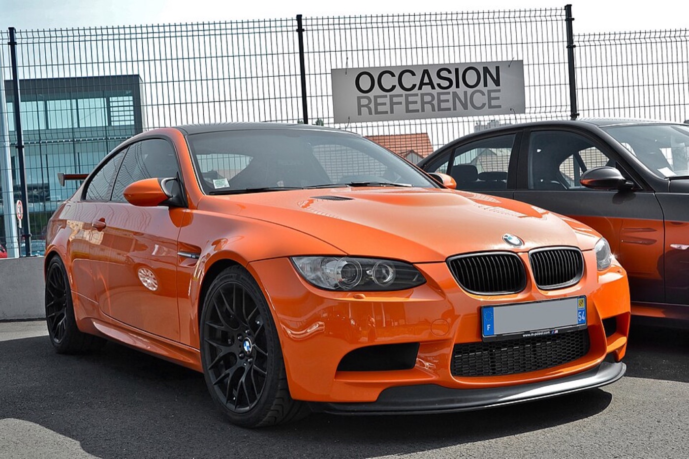 Orange BMW M3 GTS from the front three-quarter with black wheels and splitter, parked at a used-car dealership