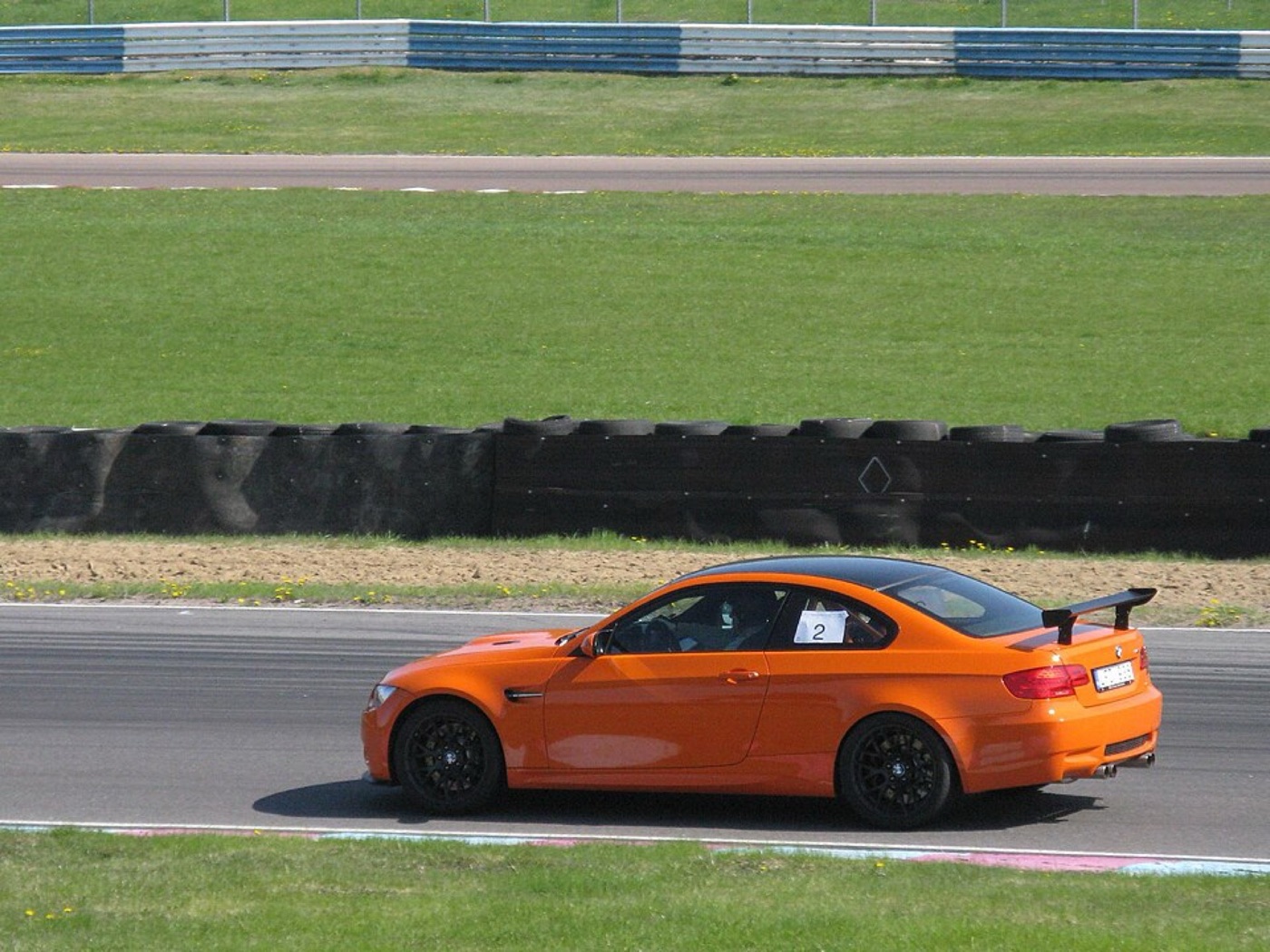 Orange BMW M3 GTS cornering on a race circuit with tyre walls behind and a number on the side window