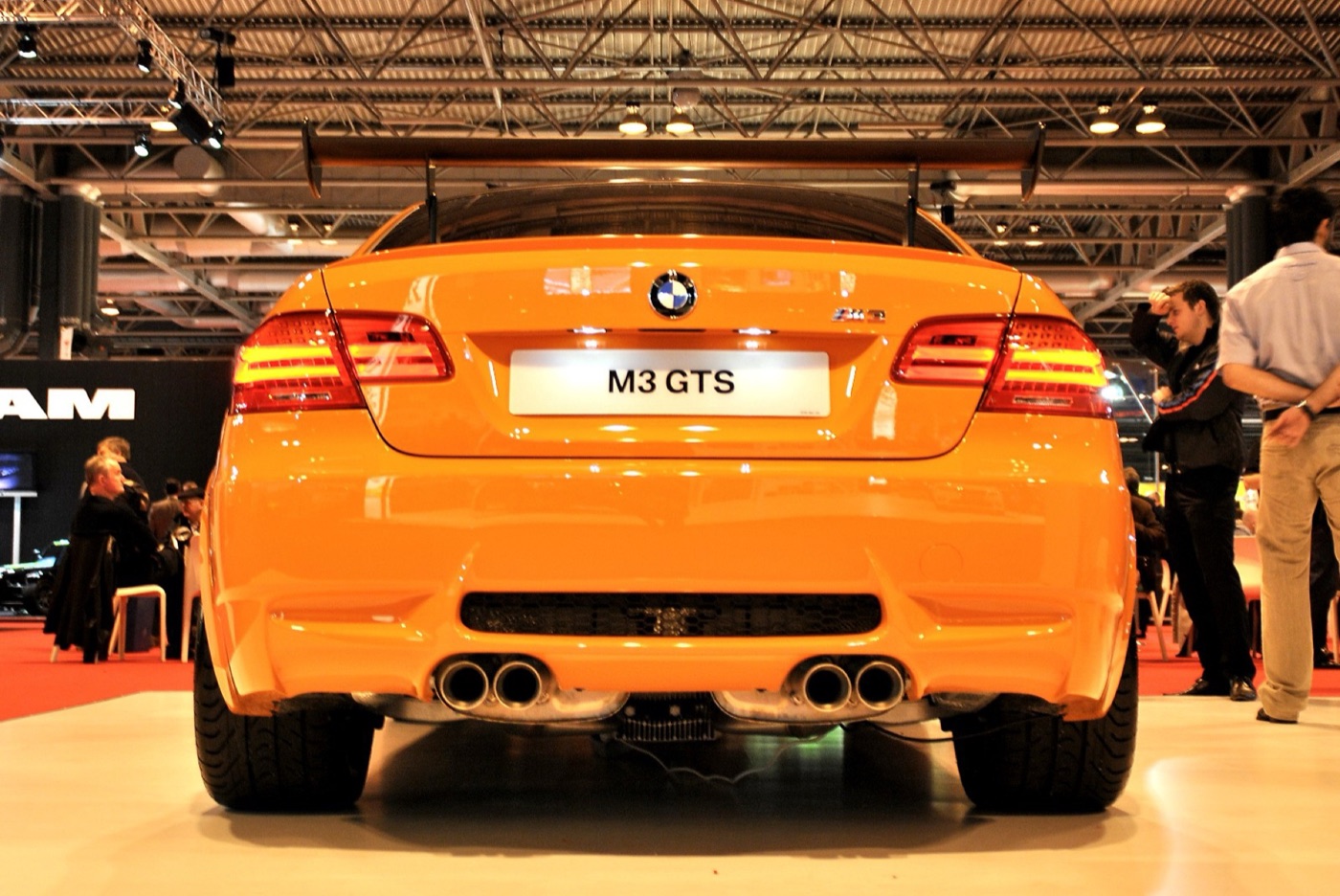 Rear view of an orange BMW M3 GTS on a show stand with an M3 GTS plate, four exhaust tips and the raised rear wing