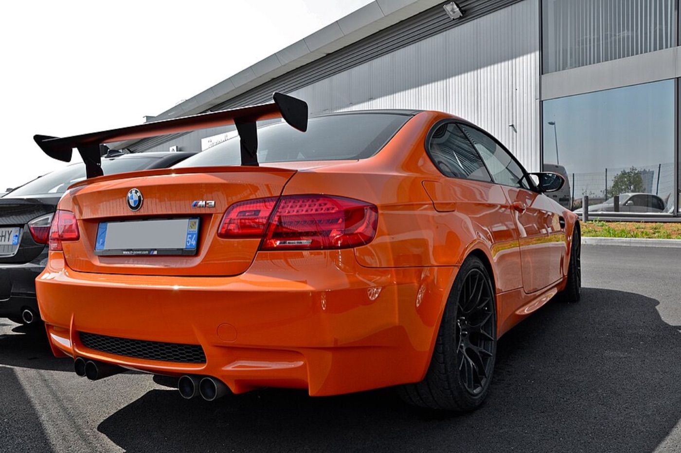 Orange BMW M3 GTS from the rear three-quarter, showing the tall adjustable rear wing and black wheels, parked outside a dealership