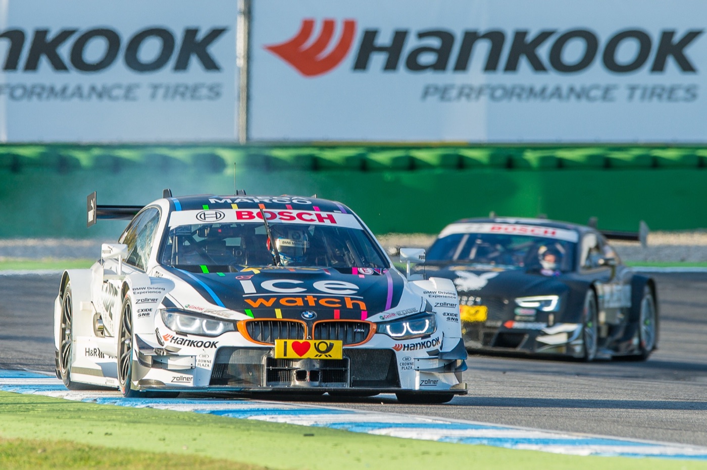 White and black Ice-Watch BMW M4 DTM racing car on a kerb, followed by a black Audi RS 5 DTM