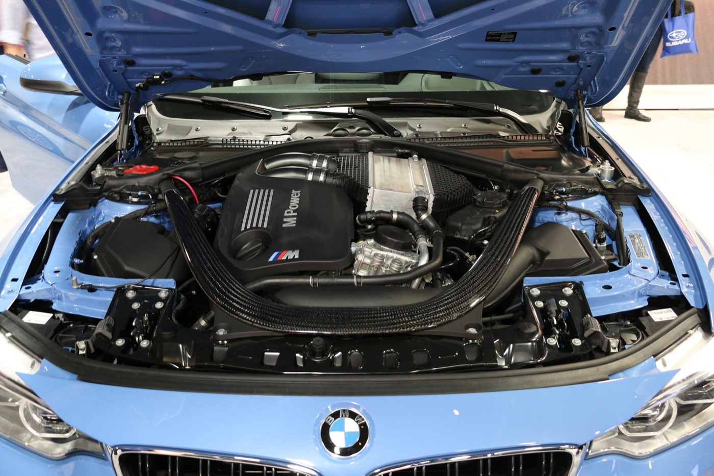 Engine bay of a blue BMW M4 with the M Power engine cover, twin intake pipes and the curved carbon-fibre strut brace