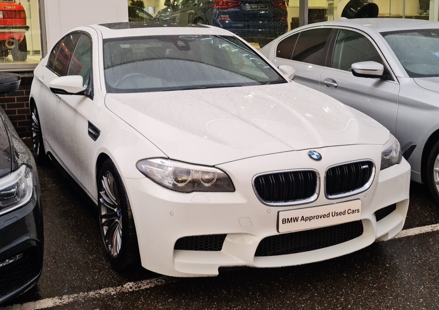 White pre-facelift BMW M5 F10 on a dealer forecourt in the rain, with a BMW Approved Used Cars plate on the front bumper