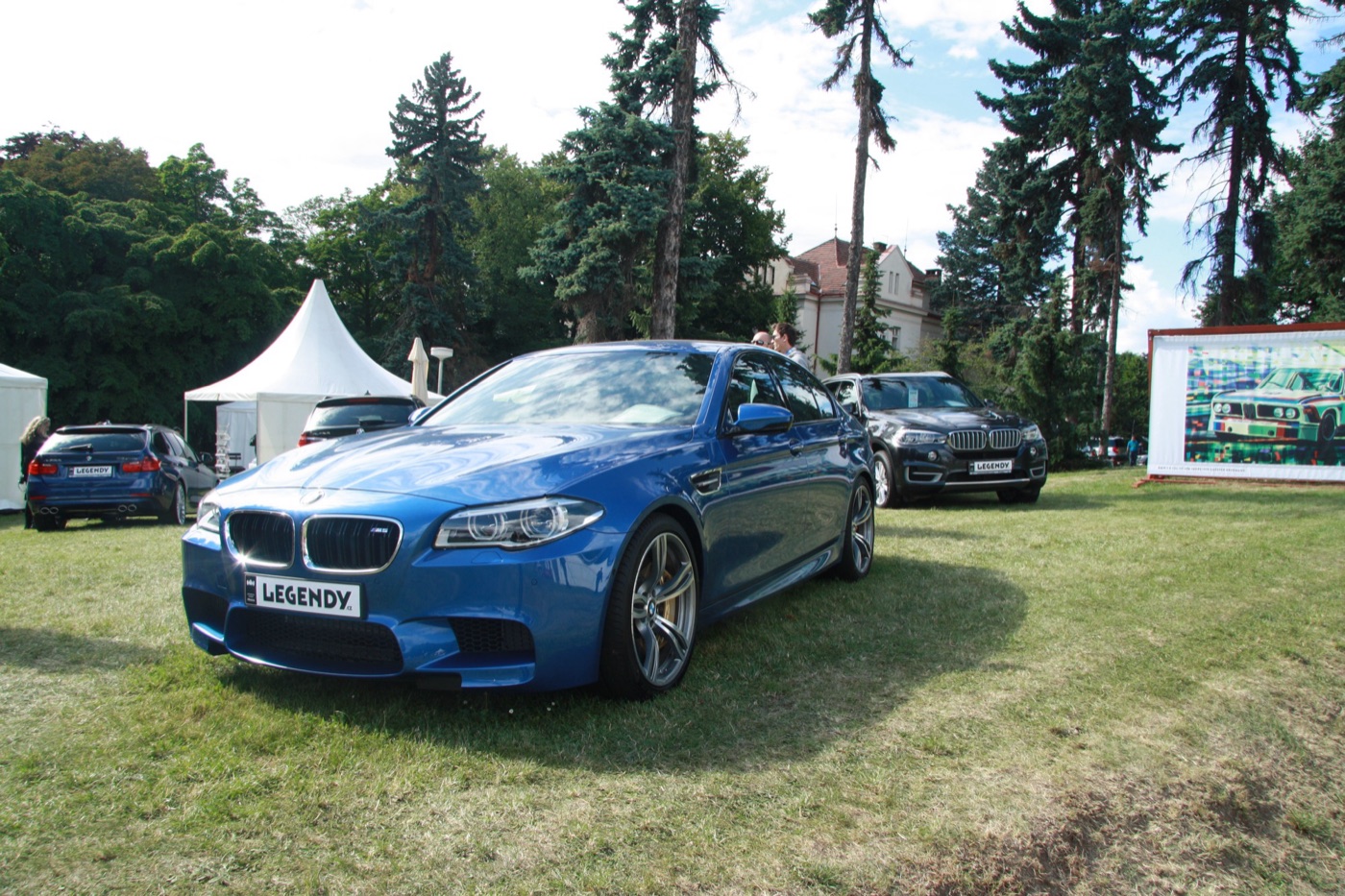 Blue BMW M5 F10 saloon parked on grass at an outdoor car show, seen from the front three-quarter with gold brake calipers