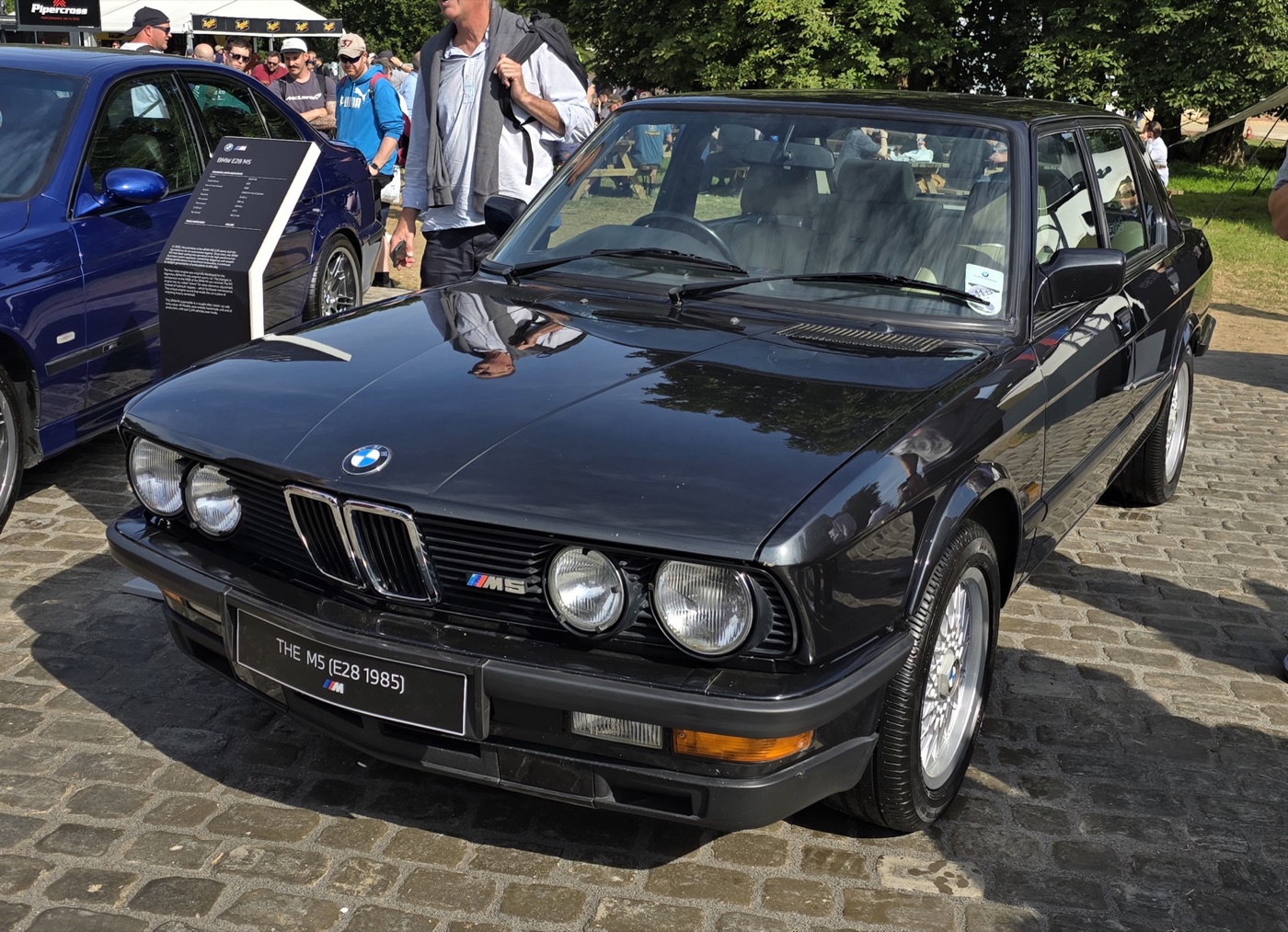 Black 1985 BMW E28 M5 saloon with four round headlamps and an M5 grille badge, parked on cobbles at a show