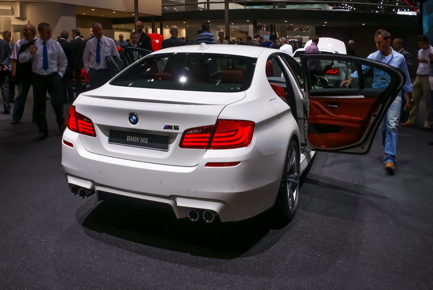 White BMW M5 F10 saloon from the rear with its rear door open to show a red leather interior, on a crowded motor show stand