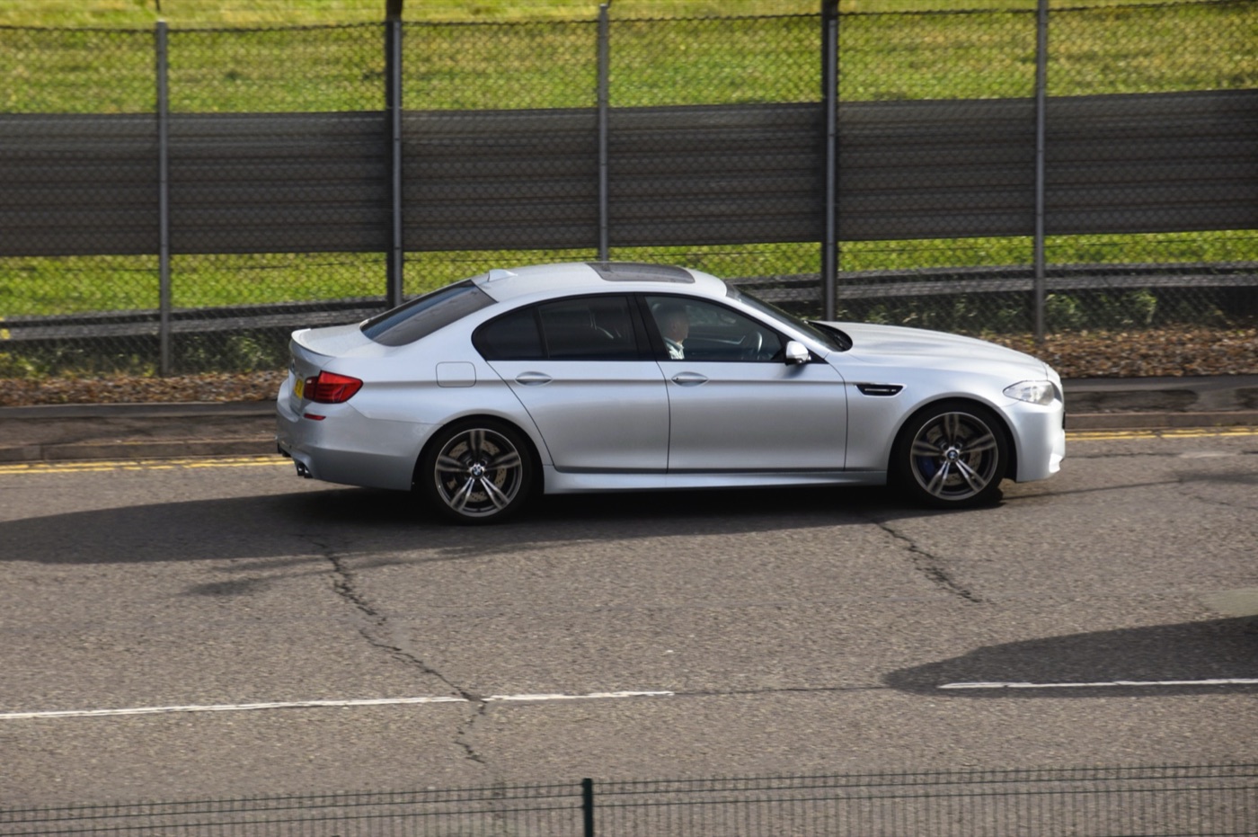 Silver BMW M5 F10 driving past a fenced verge, seen side-on with double-spoke wheels and blue front brake calipers