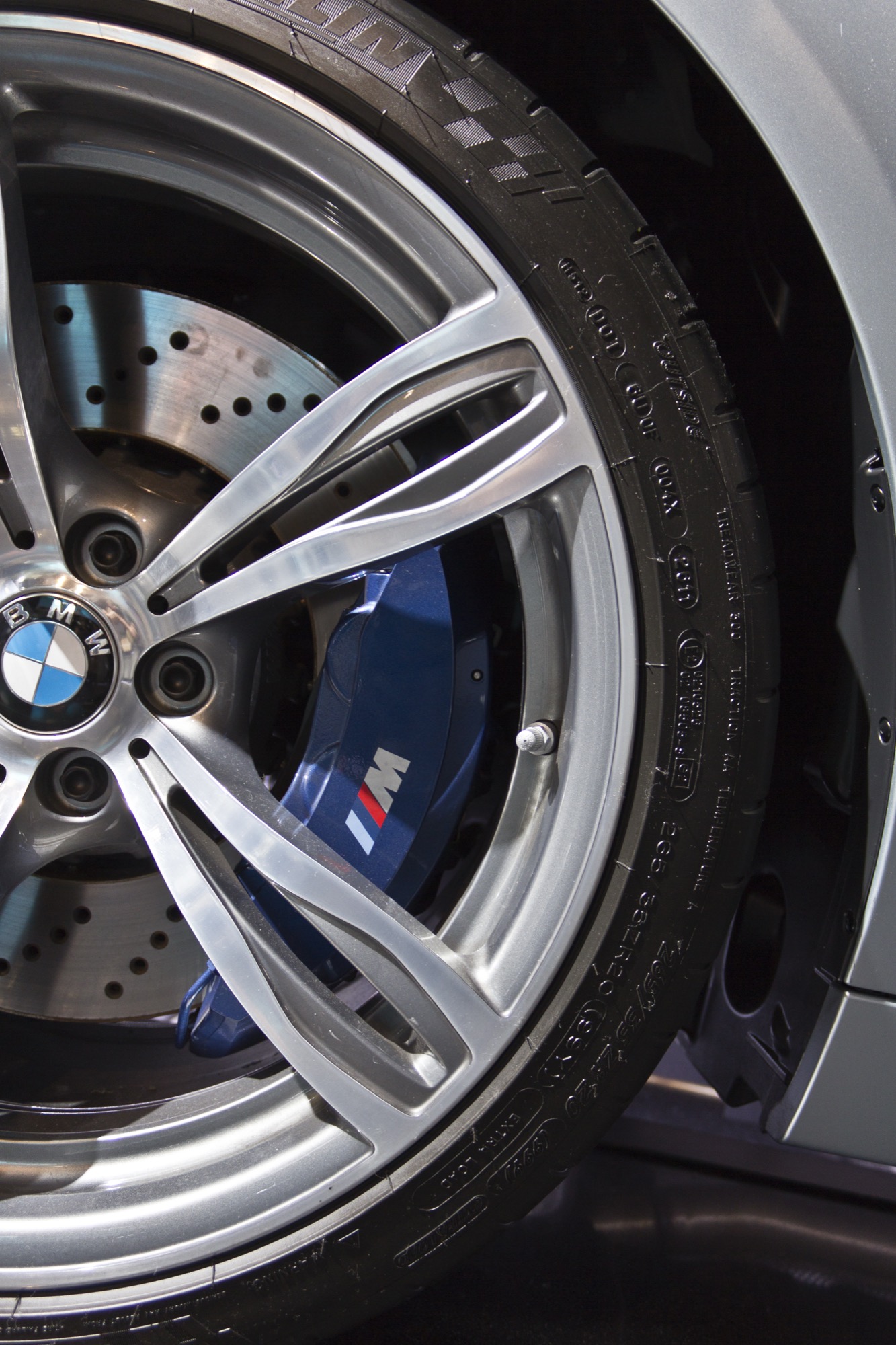 Close-up of a BMW M5 F10 20-inch double-spoke wheel showing a blue six-piston brake caliper with M logo and a cross-drilled disc