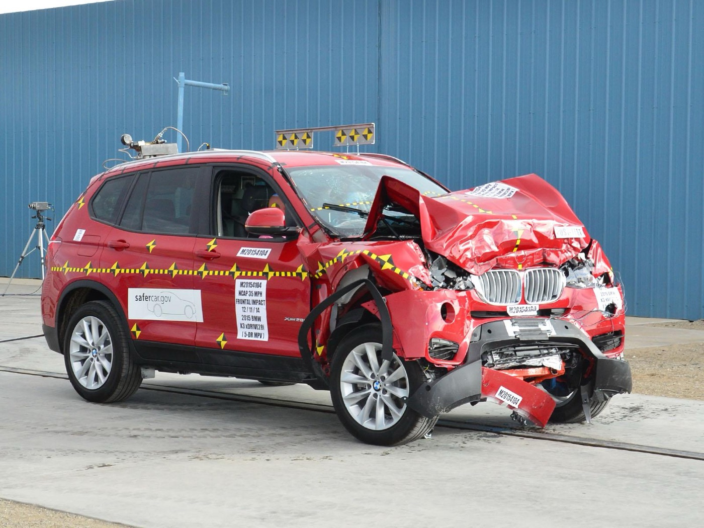 Red facelifted BMW X3 xDrive28i with its front end crushed after a full-width frontal crash test, beside a blue test-centre wall