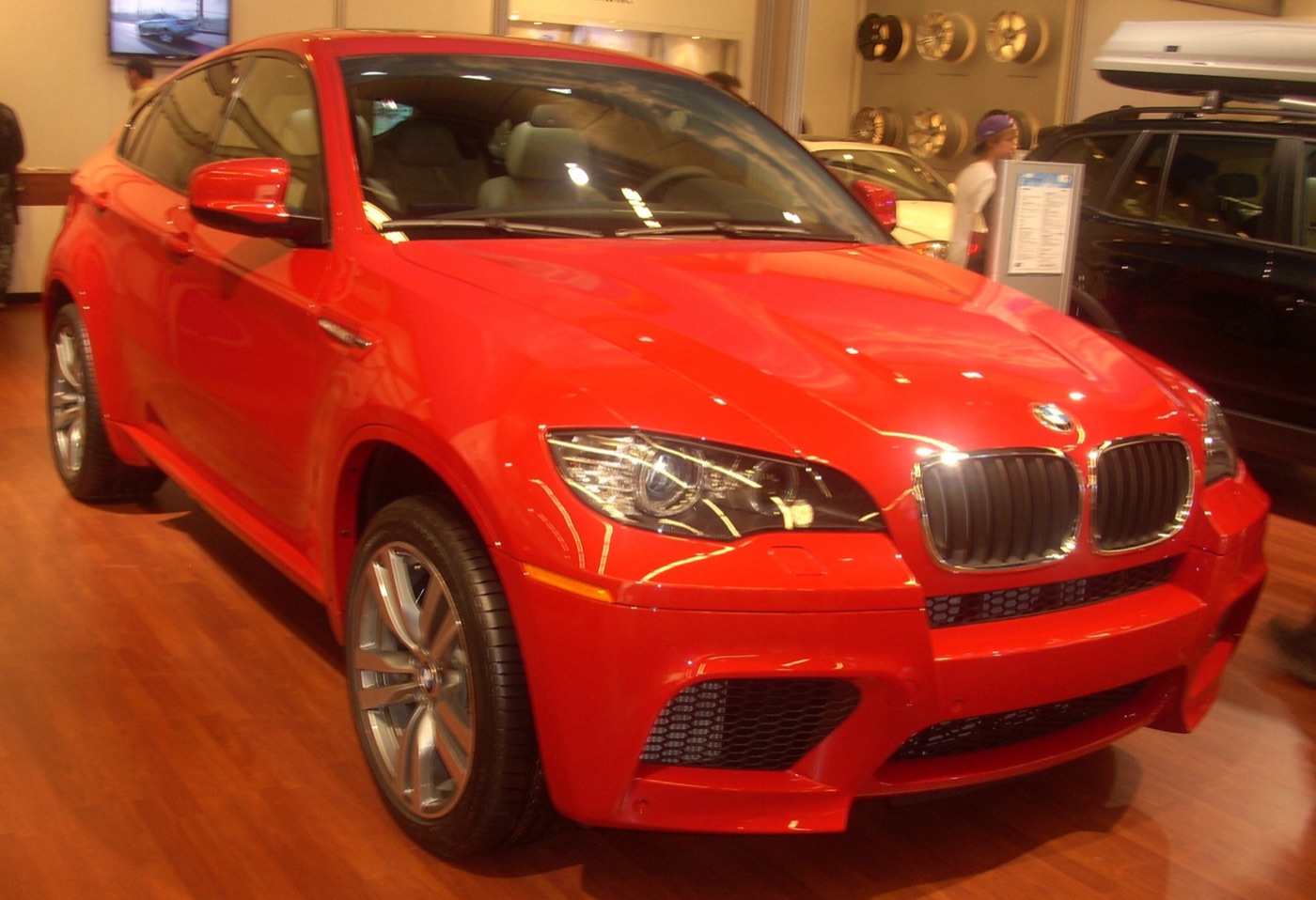 Red BMW X6 M on a motor show stand, showing its deeper front bumper with mesh air intakes