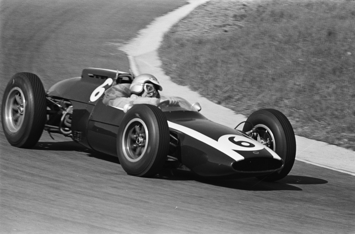 Bruce McLaren in a white helmet and goggles cornering a dark Cooper Formula One car numbered 6 on the dunes circuit