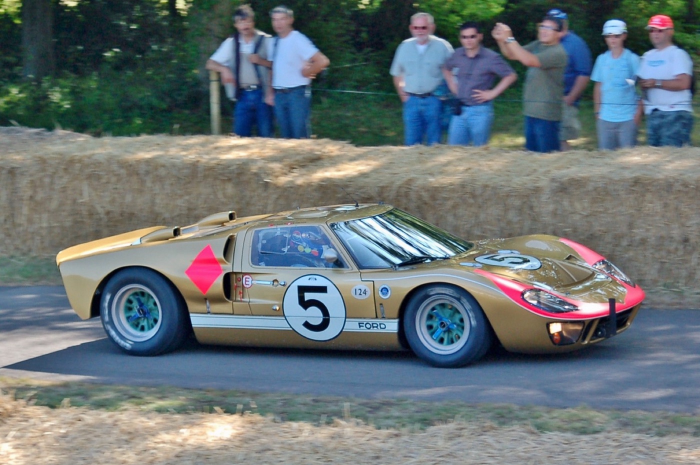 Gold Ford GT40 Mk II with a pink nose and number 5 being driven past straw bales at Goodwood