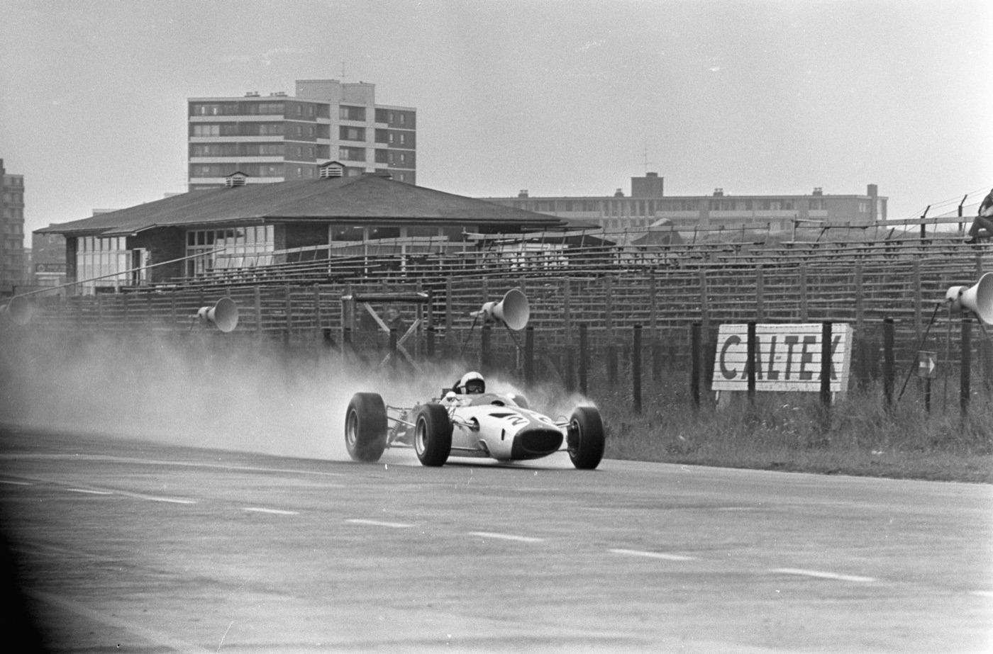 White McLaren Formula One car throwing up spray on a wet straight in front of empty wooden stands and a Caltex sign