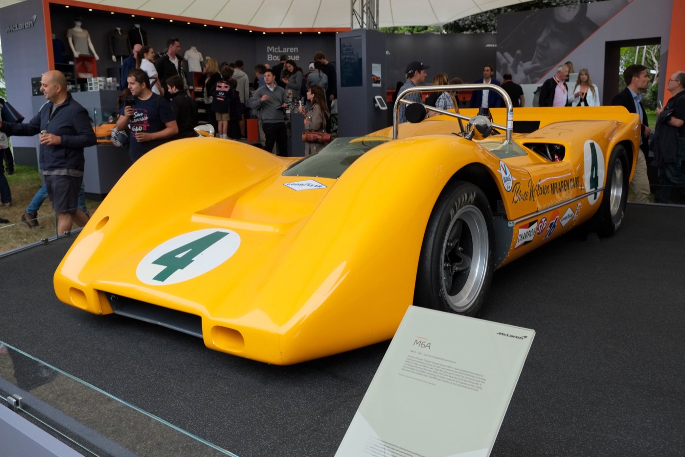 Orange McLaren M6A Can-Am car with a green number 4 and Bruce McLaren’s name on the side, displayed on a stand