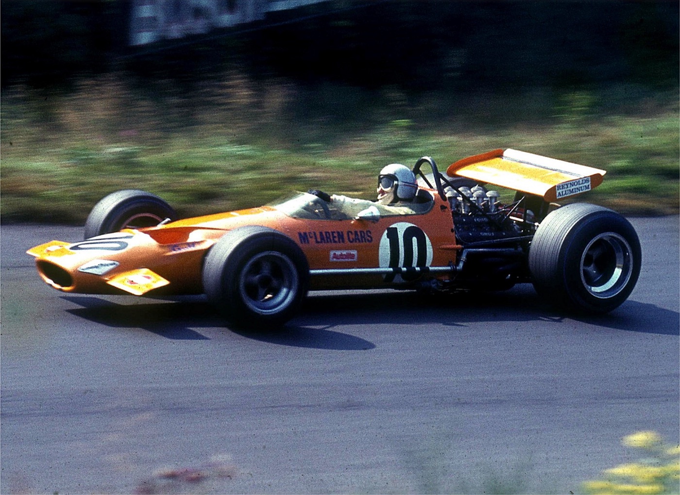 Bruce McLaren in a white helmet and goggles driving an orange McLaren Formula One car numbered 10 with a high rear wing