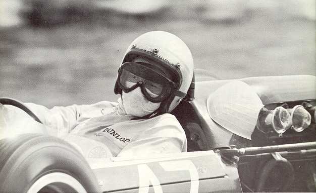 Close view of Bruce McLaren in a white helmet, goggles and face mask at the wheel of his Cooper