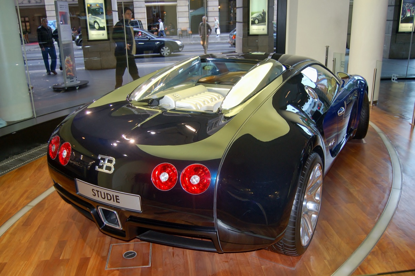 Rear three-quarter view of a dark blue and black Bugatti Veyron 16.4 on a turntable in a city showroom, its engine visible under the rear deck