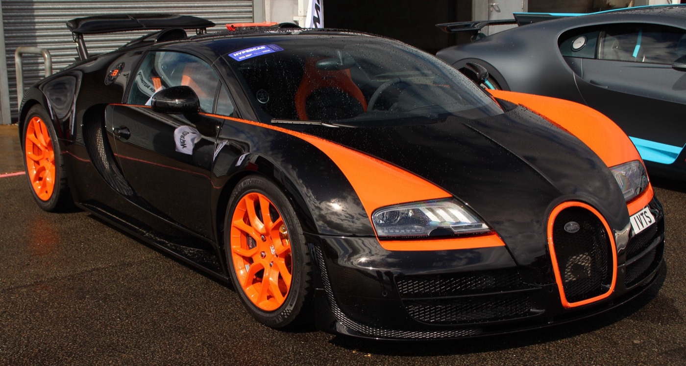 Black and orange Bugatti Veyron Grand Sport Vitesse with orange wheels, parked in a wet paddock beside other Bugattis