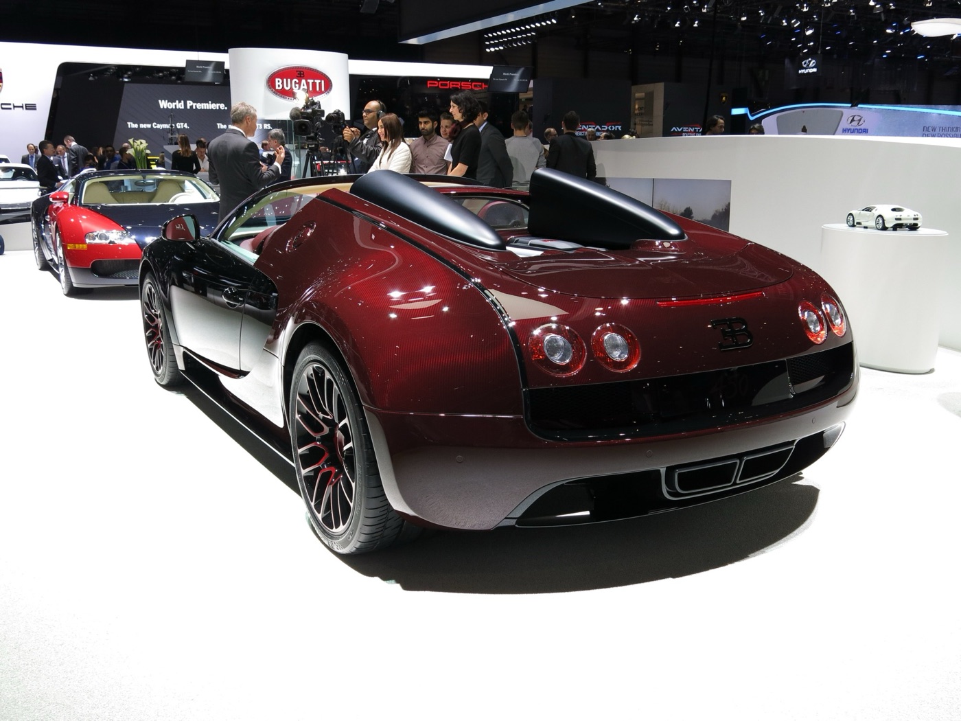 Rear view of a dark red and black Bugatti Veyron Grand Sport Vitesse on a white show stand, with a red and black Veyron coupé behind it