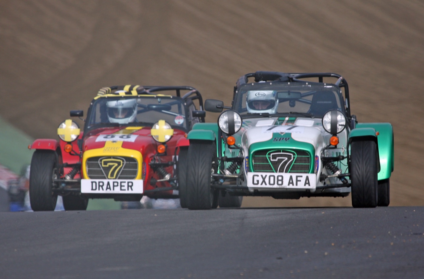 Two Caterham Sevens, one red and yellow, one green and white, racing side by side towards the camera