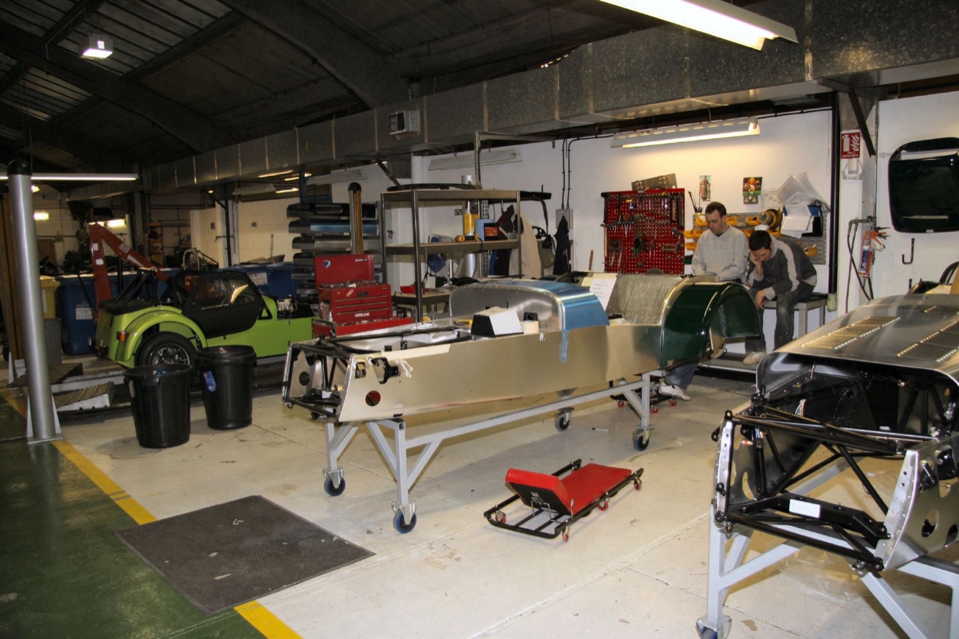 Caterham Seven chassis in bare aluminium on wheeled build stands inside the Kennet Road factory, with a finished lime-green Seven behind
