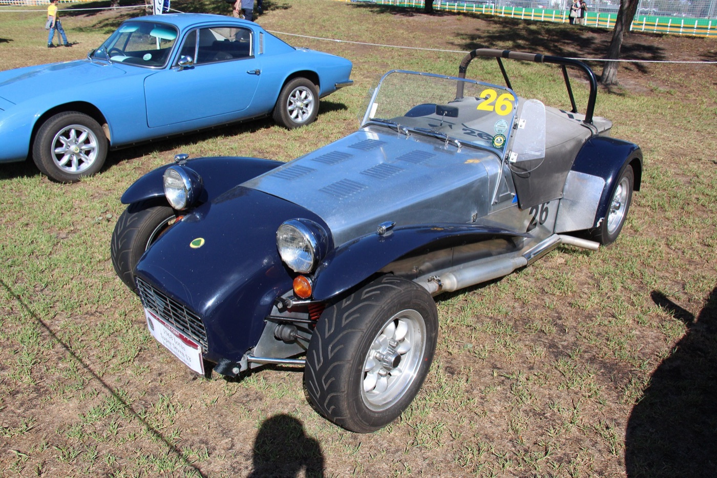 Dark blue and bare aluminium 1968 Lotus Super Seven Series 3 race car parked on grass beside a blue Lotus Elan coupé