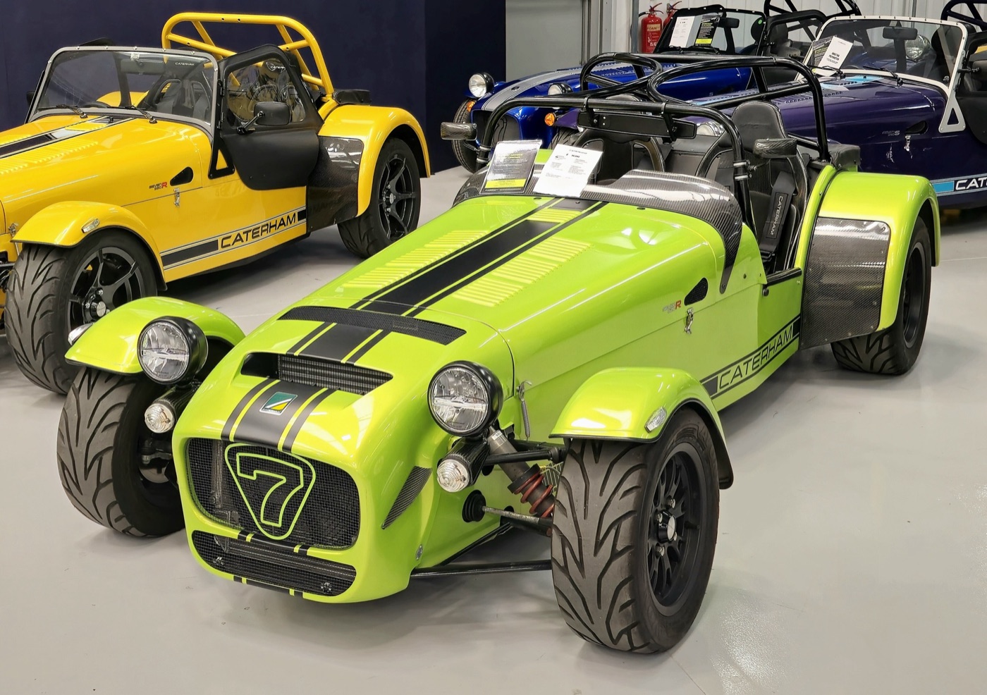 Lime green Caterham Seven 620R with black stripes and carbon-fibre wings in a showroom, a yellow Seven beside it