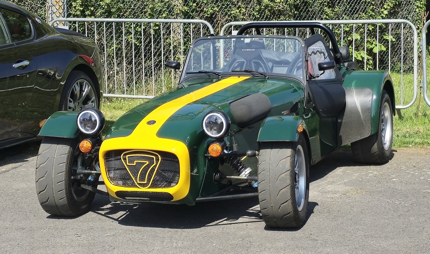 British racing green Caterham Seven with a yellow nose and centre stripe, parked by a fence